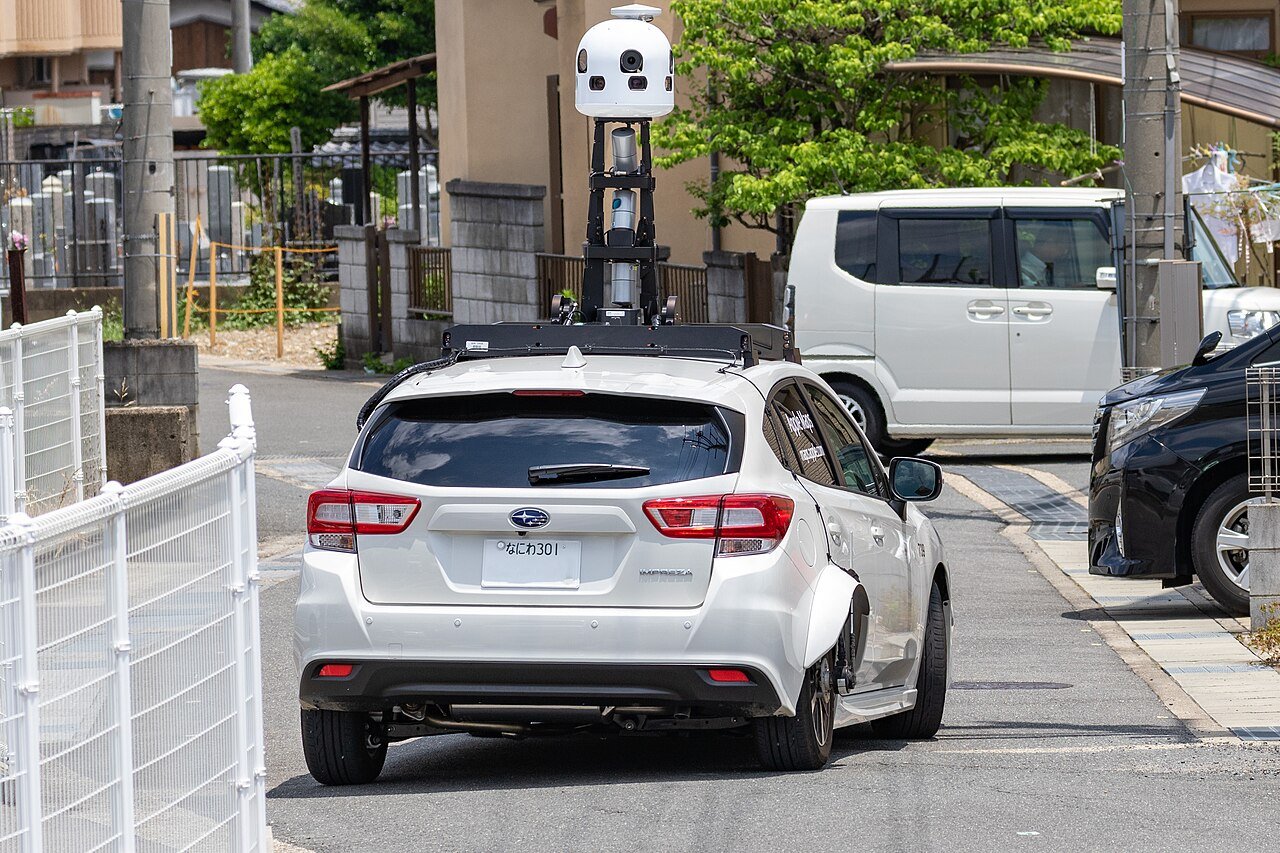 An Apple Maps survey car with a roof-mounted camera and LiDAR mast driving a narrow Japanese street