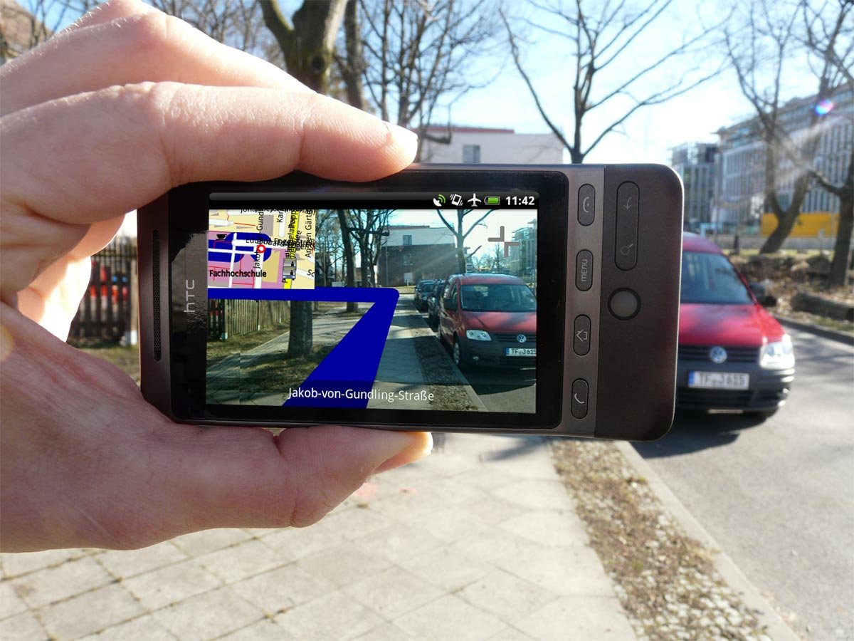 A hand holding a phone whose screen shows an AR navigation route ribbon drawn over the live camera view of the same street