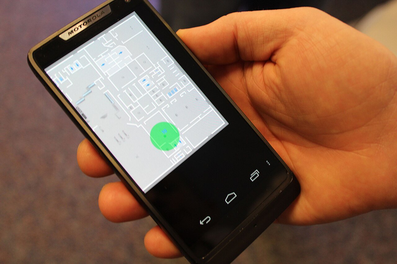 A phone showing an indoor floor plan with a green position blob marking the user's location
