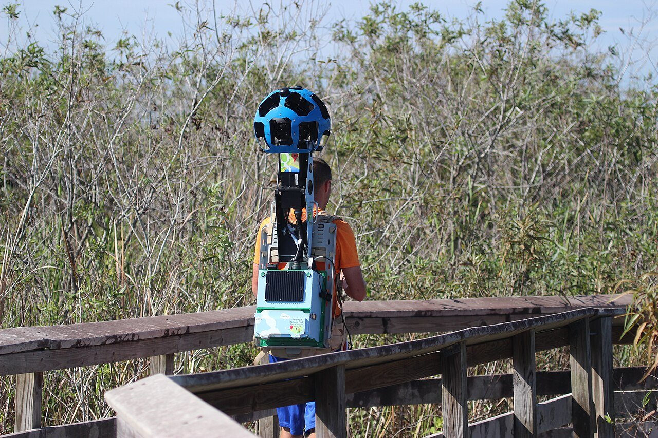 A person walking a boardwalk wearing the Google Street View Trekker backpack with its spherical camera head